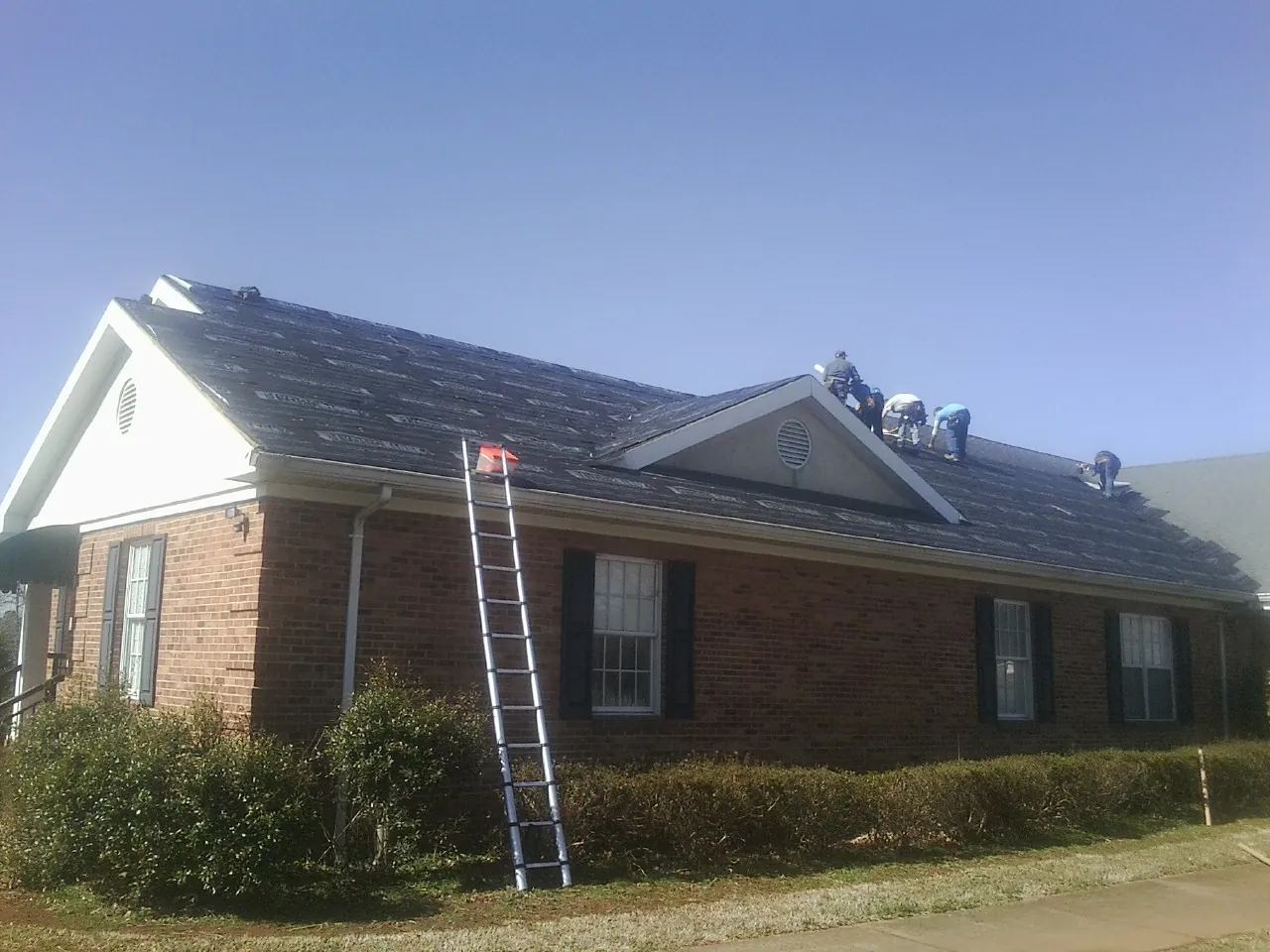 Crew tearing shingles off a brick ranch roof with a ladder set — Metal Roof Repair work by our crew in River Grove