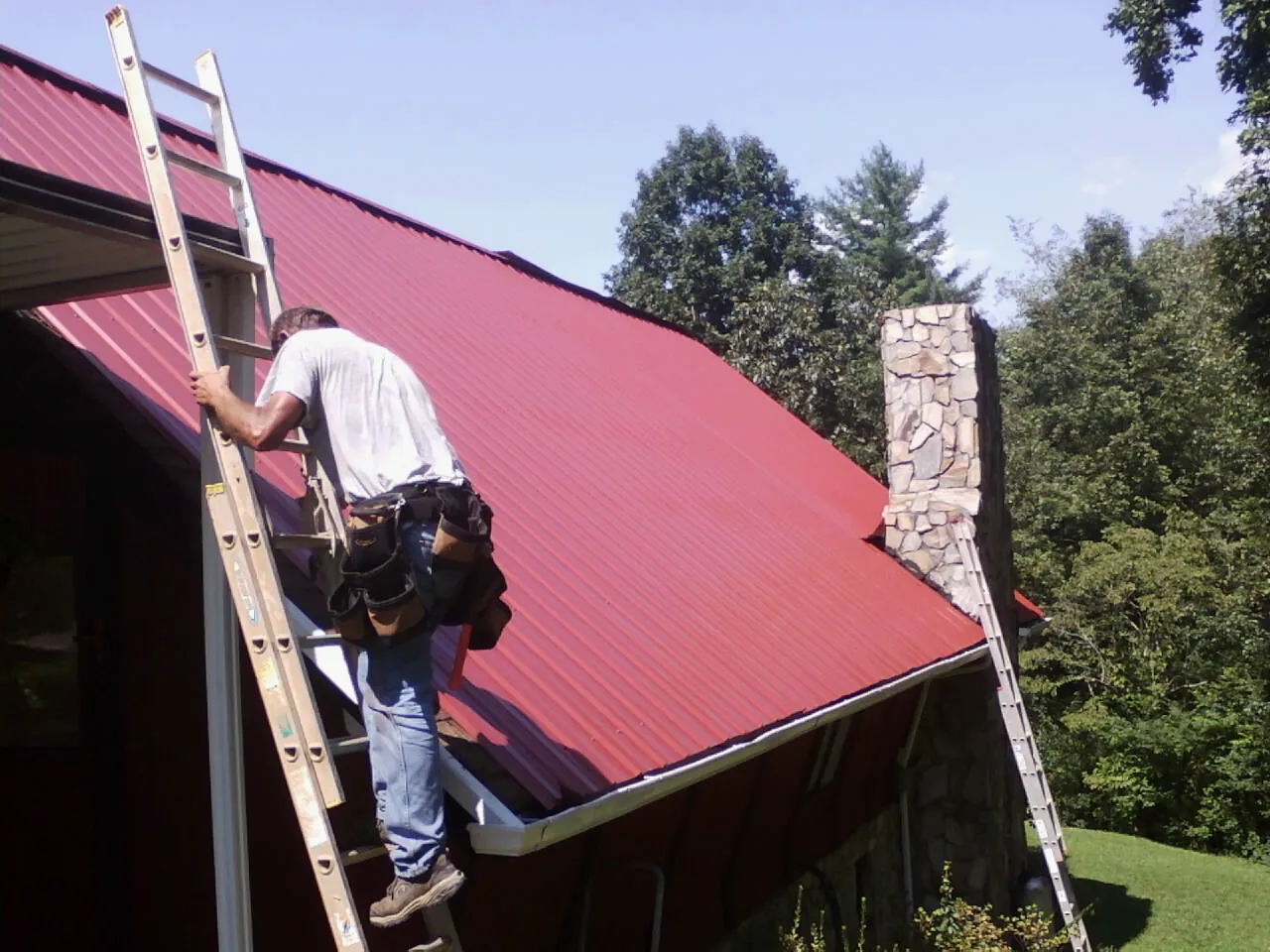 Roofer on a ladder beside a red metal roof and stone chimney — Soffit & Fascia Repair by Roofing Brothers in River Grove